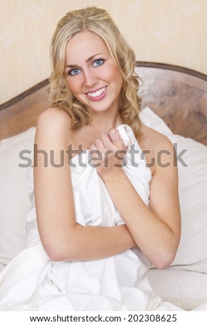 A beautiful blond haired blue eyed young woman naked and wrapped in white sheets on her bed