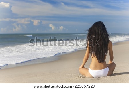 A sexy young brunette woman or girl wearing a white bikini sitting on a deserted tropical beach with a blue sky