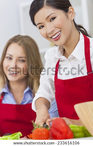 Beautiful happy young Asian Chinese woman or girl and her friend or work colleague wearing a red apron cutting & preparing fresh vegetable salad food in a kitchen