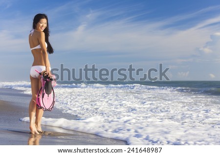 Rear view of beautiful young Asian Chinese woman in bikini with snorkel, mask & flippers on a deserted beach with blue sky
