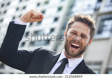 A young successful man, male executive businessman arms raised celebrating cheering shouting in front of a high rise office block in a modern city