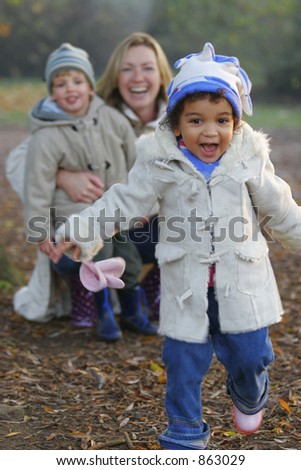 A beautiful mixed race girl runs towards the camera laughing while over her shoulder you see a young mother with her son.