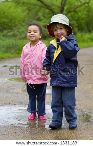 A little boy and girl wearing wellington boots holding hands and sharing a puddle in a park