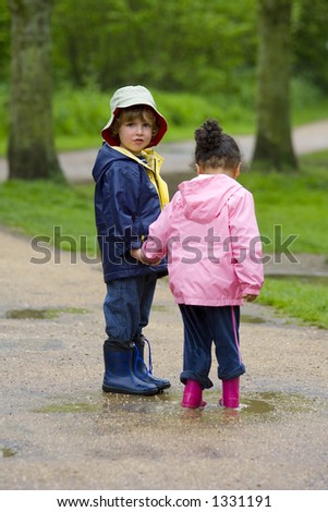 A little boy and girl wearing wellington boots, holding hands and sharing a puddle in a park