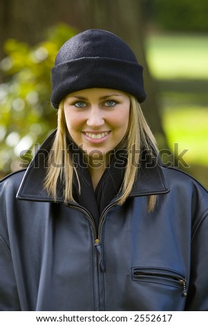 A beautiful young blonde woman in a black beanie hat and leather jacket smiles enigmatically while backlit by autumnal sunshine