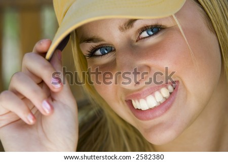 A beautiful young lady with blonde hair and blue eyes tips her baseball cap to the camera while bathed in sunshine.