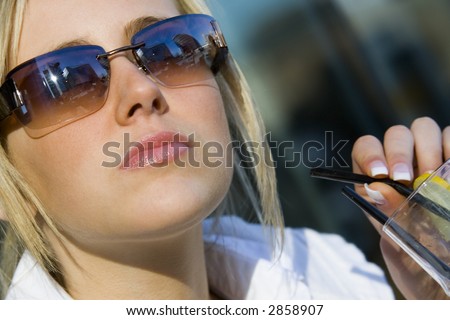 A beautiful young blonde woman relaxing and drinking a cocktail with modern hi-tech buildings reflected in her sunglasses