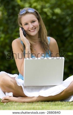 A beautiful young woman sitting cross legged in a grassy sunlit setting, working on her laptop and chatting on her mobile phone