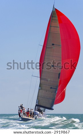 A fully crewed racing yacht with a red spinnaker catching the wind and leaving a big wake