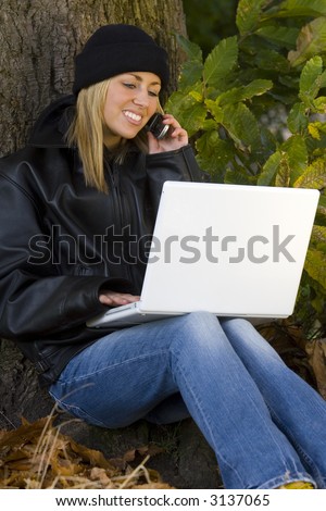 A beautiful young blond woman using a mobile phone and laptop while sitting at the base of the tree surrounded by leaves
