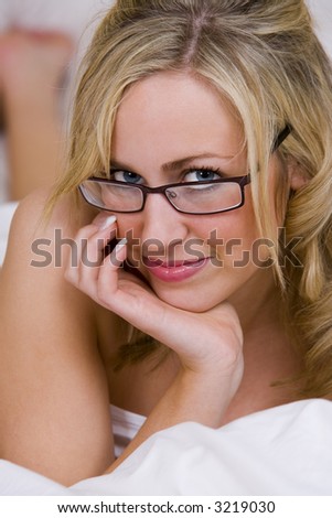 A beautiful young blond woman looking naked in bed except for white bed sheets and glasses