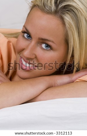 A beautiful young blond woman with stunning blue eyes laying on white bed sheets and smiling for the camera.