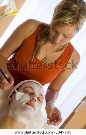A beautiful dark haired woman receives a face mask treatment from a young blond beautician