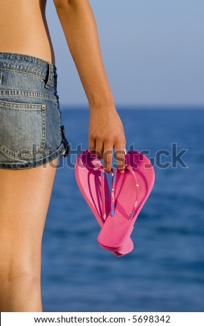Rear view of a beautiful woman standing in front of a deep blue sea carrying bright pink flip-flops
