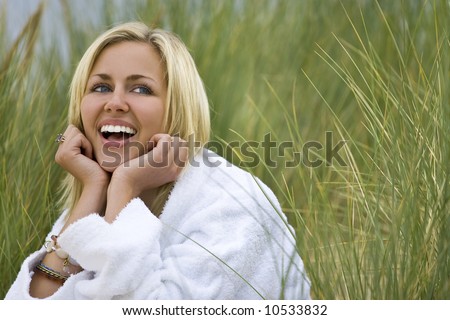 A beautiful blond haired blue eyed model wearing a white toweling robe sits laughing amid tall grass