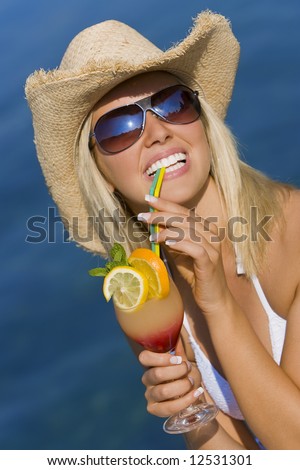 A beautiful blond haired model wearing a white bikini laughing and drinking a cocktail in front of a beautiful blue sea
