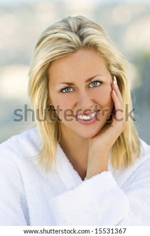 A beautiful blond haired blue eyed model wearing a white toweling robe sits relaxing outside