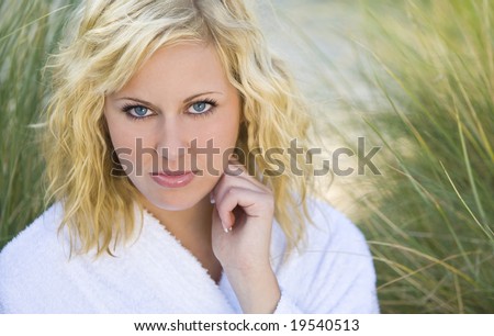 A beautiful blond haired blue eyed model wearing a white toweling robe sits amid tall grass