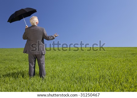 Business concept shot of a businessman standing in a green field under a bright blue sky holding and umbrella and looking towards the light. Shot on location.