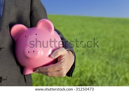 Green investment concept shot of a man in a suit holding a big pink piggy bank in a green field with a bright blue sky. Shot on location with the focus on the piggy bank in the foreground.