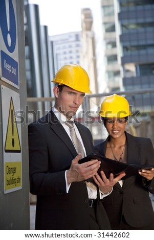 Two executives, one man and one women, wearing hard hats review plans in a modern city environment. The focus is primarily on the man in the foreground.