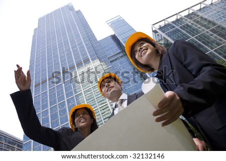 A group of three executives, one man and two women, wearing hard hats review architectural plans in a modern city environment.