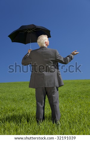 Business concept shot of a businessman standing in a green field under a bright blue sky holding and umbrella and looking towards the light. Shot on location.