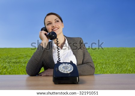 Business concept shot of a beautiful young woman sitting at a desk using an old fashioned phone in a green field with a bright blue sky. Shot on location.
