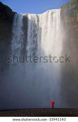 A woman dressed in a red jacket carrying a red umbrella standing at the base of a waterfall. Shot on location at Skogafoss in Iceland.