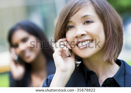 A beautiful young oriental woman with a wonderful smile chatting on her cell phone with Indian Asian woman on her phone out of focus behind her.