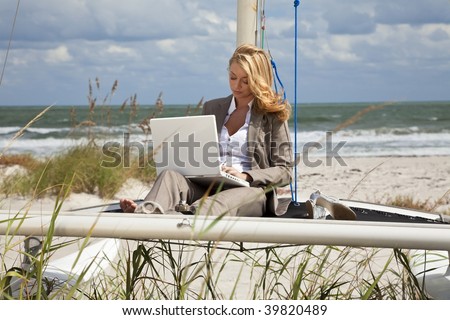 A beautiful young woman in a smart suit sitting barefoot on the deck of a small catamaran sailing boat using her laptop computer with the beach and sea behind her