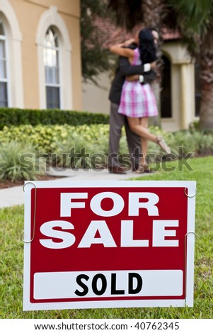 House For Sale and Sold sign with African American couple celebrating the purchase of a house out of focus behind the sign.