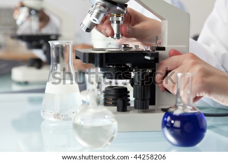A medical or scientific researcher using a microscope in a laboratory environment with flasks and medical equipment.