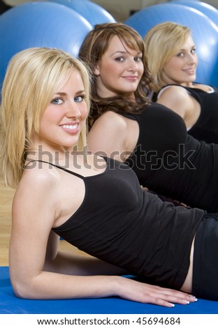 Three beautiful young women, two blond, one brunette, working out on on mats doing aerobic sit ups at the gym. The focus is on the blond girl with blue eyes in the foreground.