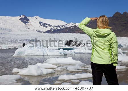 Environmental picture of woman hiker dressed in green jacket looking at the effects of global warming climate change on a melting glacier with icebergs floating into a glacial pool.