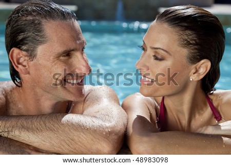 Close up portrait of a beautiful happy man and woman couple looking at each other and resting on their hands at the side of a sun bathed swimming pool smiling with perfect teeth.