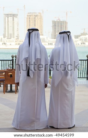 Two anonymous Arab men in traditional white clothing of dish dash and shemagh looking at the construction of new modern skyscrapers on the horizon