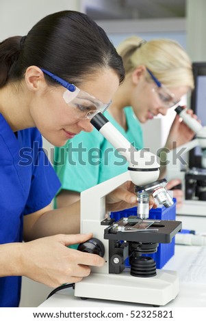 A blond female medical or scientific researcher or doctor using her microscope in a laboratory with her  colleague out of focus behind her.