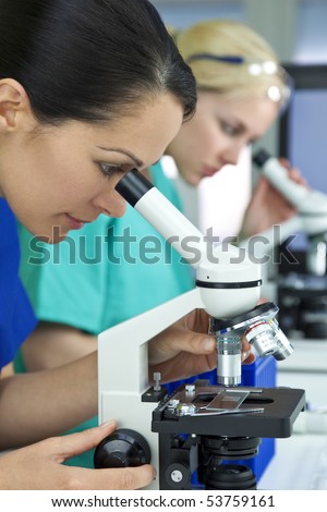 A female medical or scientific researcher or doctor using her microscope in a laboratory with her blond colleague out of focus behind her.