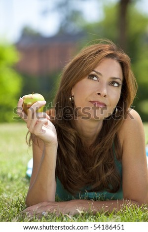 A beautiful woman in her thirties laying down outside on grass holding or eating an apple looking up and thinking