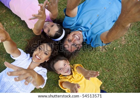 An attractive smiling family of mother, father, and two children laying down on grass outside having fun with arms and hands outstrectched to the camera
