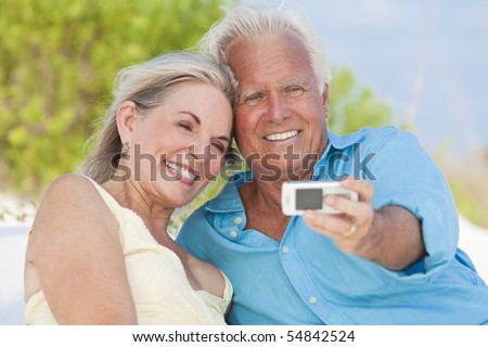 Happy senior man and woman couple laughing and taking photographs with a cell phone on a tropical beach with bright clear blue sky