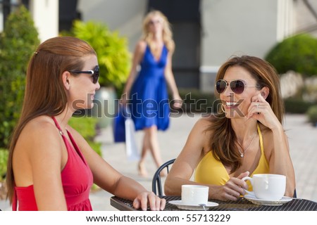 Two beautiful and sophisticated young women friends wearing sunglasses and having coffee around a modern city cafe table With their friend arriving with shopping bags in the background