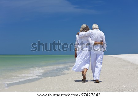 Rear view of a senior man and woman couple walking arms around each other on a deserted tropical beach with bright clear blue sky
