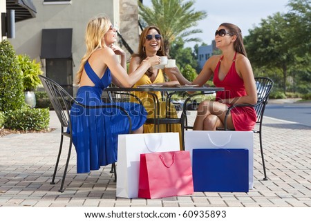 Three beautiful and sophisticated young women friends wearing sunglasses and having coffee around a modern city cafe table surrounded by shopping bags