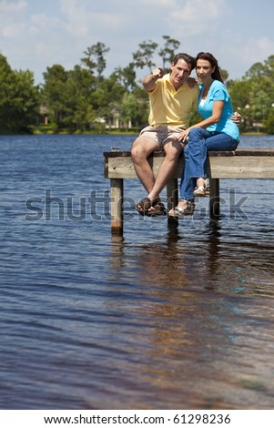 Portrait shot of an attractive and happy middle aged man and woman couple in their thirties, sitting together on a pier by a lake and pointing.
