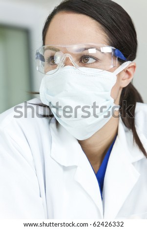 An attractive female doctor or scientist wearing a white coat and surgical face mask and safety glasses in a hospital.