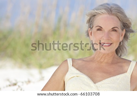 An attractive elegant senior woman sitting on a white sand beach with grass and a blue sky behind her.