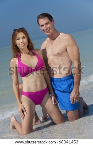 Happy fit and healthy man and woman middle aged couple in swimming trunks and bikini kneeling on a deserted beach with bright clear blue sky