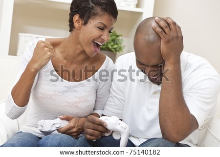 African American couple, man and woman, having fun playing video console games together. The woman has just beaten the man, she is celebrating, he is laughing.